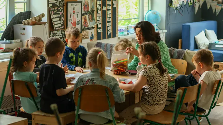 Eine Erzieherin sitzt mit einer Gruppe Kinder an einem Tisch im Gruppenraum
