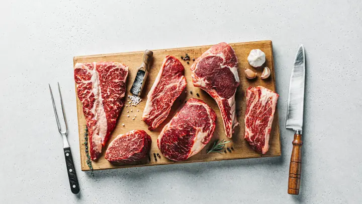 Rohe Rindersteaks auf einem Holzbrett mit Messer und Gabel vor hellem Hintergrund