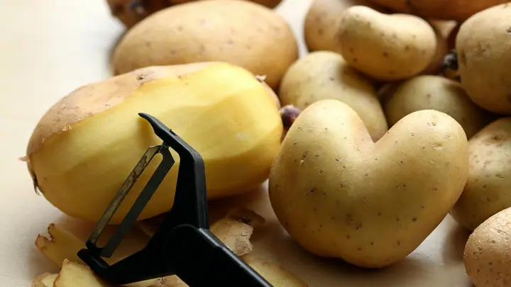 Rohe Kartoffeln auf einem Holztisch, eine davon halb geschält, daneben ein Sparschäler