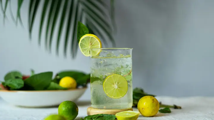 Glas Limonade mit Limettenscheiben und Eis, daneben Zitronen, Limetten und Blätter auf hellem Tisch