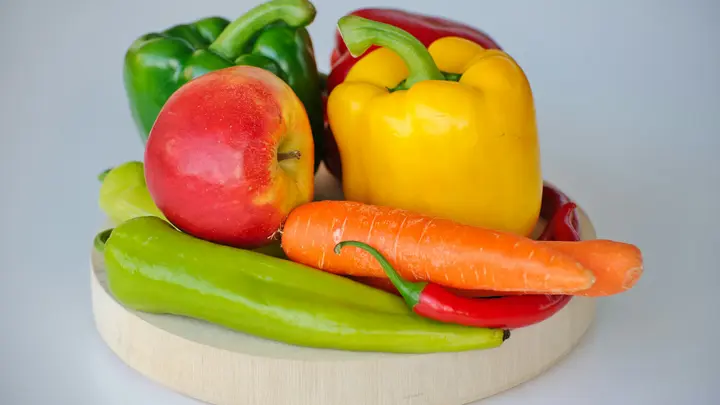 Frische Paprika, Möhren und ein Apfel auf einem hellen Holzteller