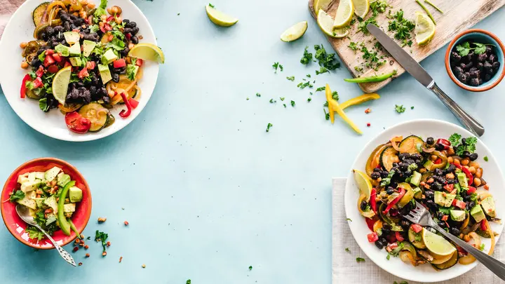 Salate mit schwarzen Bohnen, Kichererbsen, Avocado und Gemüse auf hellblauem Tisch, daneben Limetten
